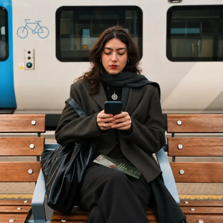 Frau sitzt mit Handy an Bahnhof.