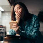 eine Frau gähnt, während sie ein Sandwich hält